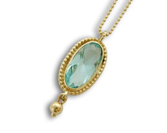 Op de afbeelding: Een gouden ketting met een lichtblauwe ovale edelsteen hanger en een kleine gouden kraal aan het einde.