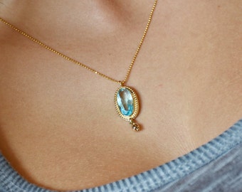 Op de afbeelding: Een gouden ketting met een lichtblauwe edelsteen hanger. De hanger is ovaalvormig en heeft een gouden rand.