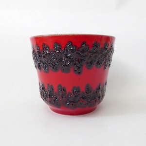 May include: A ceramic cup featuring a vibrant red glazed finish with two horizontal bands of dark, textured, porous black lava glaze. The cup has a wide, open top and a tapered base, showcasing a contrast between the smooth red and rough black.