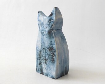 Könnte beinhalten: Eine stilisierte Katzenfigur mit einem blau-weißen Wasch-Finish. Die Oberfläche zeigt eingravierte Details eines Katzengesichts oben und ein großes Sonnenblumenmuster auf dem Körper. Das Objekt hat eine glatte, vertikale Form.
