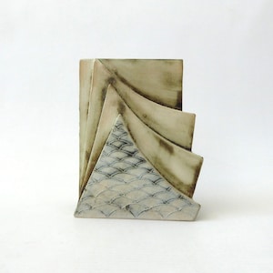 May include: A ceramic sculpture featuring four layered, fan-like panels with a cream-coloured finish and dark, weathered edges. The front-most panel displays a repeating, etched scale pattern in a soft blue-grey tone, creating a textured effect.