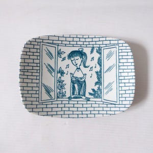 May include: A rectangular white ceramic tray featuring a blue line drawing of a person standing in an open window. The illustration depicts a brick wall background, leafy vines, musical notes, and two small birds held by the central figure.