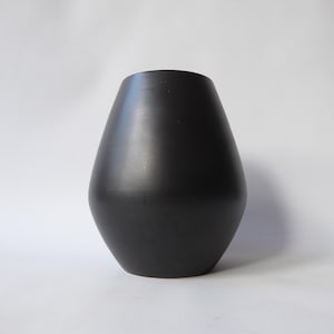 May include: A black ceramic vase with a smooth, matte finish. The vase has a rounded, teardrop shape, wider at the base and narrowing towards the top. The vase is a decorative item for the home, with a simple yet elegant design. The vase is approximately 25 cm tall.