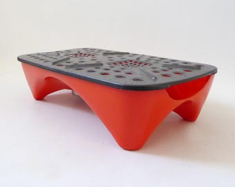 Könnte beinhalten: Ein rechteckiger Untersetzer mit einer leuchtend orangefarbenen Basis mit vier stabilen Beinen und einer abnehmbaren dunkelgrauen Deckplatte. Die Deckplatte ist mit einer perforierten Oberfläche aus zahlreichen kreisförmigen Löchern gestaltet.