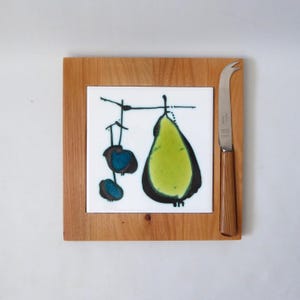 May include: A square wooden serving board featuring a central white ceramic tile with a stylised green pear and two blue circular shapes. A metal cheese knife with a matching wooden handle is positioned vertically along the right side of the board.