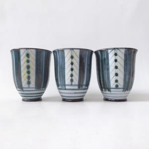 May include: Three ceramic teacups with a blue and white striped design. Each cup features a vertical pattern of yellow dots and a dark brown base. The cups are arranged side by side against a white background.