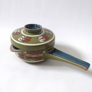 May include: A vintage ceramic hot pot with a lid. The pot is olive green with a blue handle and rim. The lid features a floral design in white, brown, and orange. The text "Ceramano HOT-POT handgemalt" is visible on the front.