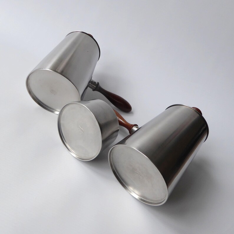 May include: Three silver pitchers with wooden handles. The pitchers are different sizes and are all made of polished metal.