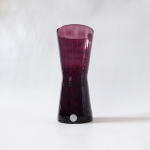May include: A deep purple glass vase with a flared rim and a tapered waist. The surface features a subtle vertical ribbed texture throughout the body, and a small circular white sticker is affixed near the base of the vessel.
