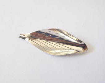 Könnte beinhalten: Eine polierte, silberfarbene Metallschale in Blattform. Das Objekt hat eine glatte, reflektierende Oberfläche mit eingravierten Linien, die die Blattadern nachahmen und von einem zentralen Grat zu den spitz zulaufenden Rändern verlaufen.