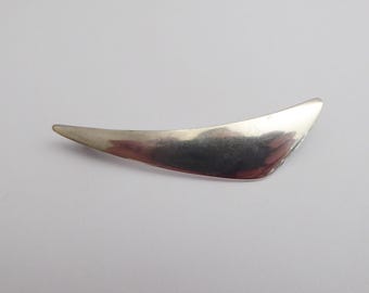 May include: A polished silver-toned metal brooch shaped like a curved, elongated crescent or blade. The surface is smooth and reflective, tapering to pointed ends on both sides, with a subtle arc that gives it a sleek, streamlined appearance.