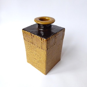 May include: A square, textured ceramic vase with a dark brown top and a golden-brown body. The vase has a circular opening at the top and a textured, vertical pattern on the sides. The vase is a decorative home decor item.
