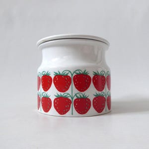 May include: A white ceramic jar with a lid, featuring a repeating pattern of bright red strawberries with green stems and leaves arranged in two horizontal rows around the cylindrical body of the container.