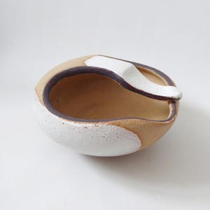 May include: A small, round, hand-formed ceramic bowl with an irregular, organic shape. The exterior features a textured, matte white glaze on the lower half, while the upper rim is accented with a smooth, dark brown band and a white ceramic handle.