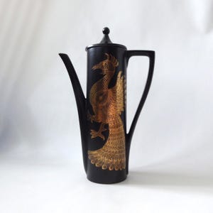 May include: A tall, matte black coffee pot featuring a slender, elongated body, a long curved spout, and a matching lid with a small spherical knob. A detailed gold-coloured illustration of a phoenix with intricate feather patterns adorns the front.