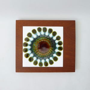 May include: A square wooden wall plaque featuring a central ceramic tile. The tile displays a circular, radial pattern with a deep brown centre, surrounded by concentric rings of olive green, teal, and white, finished with a glossy glaze.