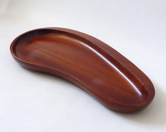 May include: A smooth, dark brown wooden tray with a distinct kidney-bean shape. The tray features a shallow, concave interior surface with a polished finish, showcasing the natural wood grain patterns throughout the curved, elongated form.