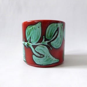 May include: A cylindrical ceramic mug featuring a glossy deep red base colour with a prominent embossed pattern of light mint green leaves and vines wrapping around the exterior surface. The design has a smooth, glazed finish throughout.