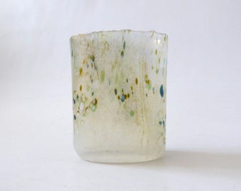 May include: A cylindrical glass tumbler with a translucent, off-white base featuring scattered, small, multi-coloured flecks in shades of blue, green, and brown. The glass has a slightly uneven, textured rim and a smooth, rounded bottom edge.
