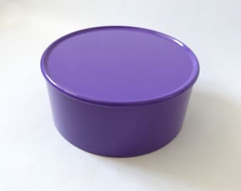 May include: A solid purple circular container with a matching flat lid. The object features a smooth, glossy finish and a simple cylindrical shape with a slightly tapered base, presenting a uniform monochromatic appearance throughout the entire item.