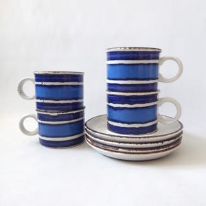 May include: A set of three ceramic cups and three matching saucers featuring horizontal bands of dark blue, light blue, and white. The cups have a cylindrical shape with a side handle, and the edges of the saucers and cups show a brown rim.