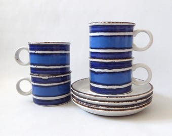 May include: A set of three ceramic cups and three matching saucers featuring horizontal bands of dark blue, light blue, and white. The cups have a cylindrical shape with a side handle, and the edges of the saucers and cups show a brown rim.