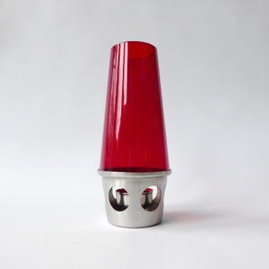May include: A red glass candle holder with a stainless steel base. The candle holder has a cylindrical shape with a flared top. The base has two circular cutouts. The candle holder is designed to hold a single candle.