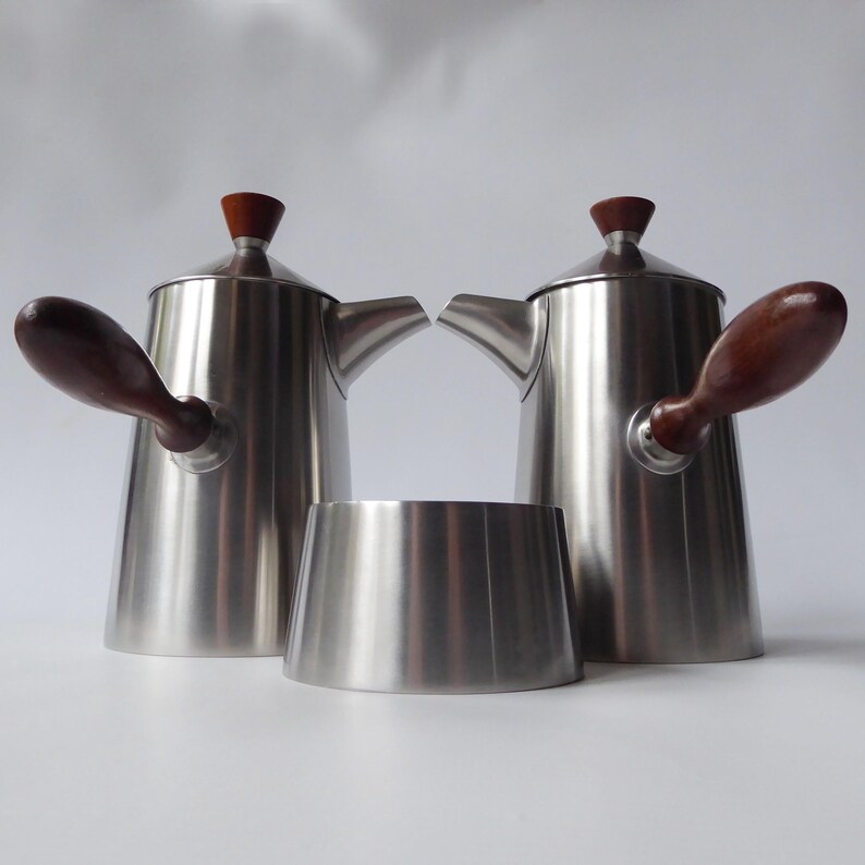 May include: A set of two silver coffee pots with wooden handles and lids. The pots are sitting on a white surface.