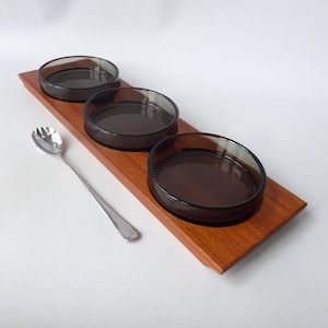 May include: A wooden serving tray with three round, dark brown glass bowls. A silver serving spoon rests on the left. The tray is rectangular with a rich, reddish-brown finish, ideal for serving appetizers or snacks.
