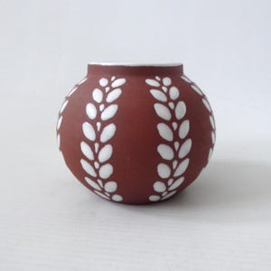 May include: A spherical, terracotta-coloured ceramic vase with a white rim and white leaf-like patterns. The vase has a smooth surface and is suitable for displaying flowers or as a decorative accent piece. The design is symmetrical and the vase is centered.