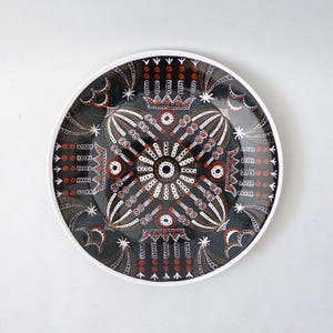 May include: A round ceramic plate featuring a dark black background with a symmetrical, intricate pattern in white, red, and brown. The design includes central circular motifs, radiating lines, star shapes, and crown-like elements.