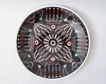 Könnte beinhalten: Ein runder Keramikteller mit schwarzem Hintergrund und einem symmetrischen, komplexen Muster in Weiß, Rot und Braun. Das Design umfasst zentrale kreisförmige Motive, strahlenförmige Linien, Sternformen und kronenartige Elemente.