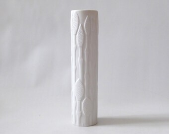 May include: A tall, cylindrical white ceramic vase featuring a textured relief pattern of vertical stems and oval-shaped seed pods. The surface has a matte finish, and the design wraps around the entire exterior of the vessel, creating a tactile, organic appearance.