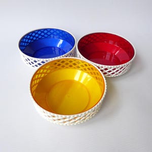 May include: Three colourful plastic bowls. Each bowl has a white lattice-style rim and a solid interior. The bowls are in primary colours: blue, red, and yellow. The bowls are round and appear to be empty.