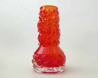 Könnte beinhalten: Eine leuchtend orangefarbene Glasvase mit einer strukturierten Oberfläche, die erhabene kreisförmige und abstrakte geometrische Muster aufweist. Die Vase hat einen breiten, runden Boden, der sich zu einem schmaleren, zylindrischen Hals verjüngt.