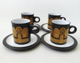 Könnte beinhalten: Ein Set aus vier schwarzen Keramiktassen und Untertassen mit weißem Innenraum und einem sich wiederholenden goldfarbenen geometrischen Muster auf der Außenseite jeder Tasse. Jede Tasse hat einen runden Henkel und steht auf einer passenden Untertasse.