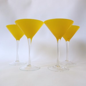 May include: Four yellow martini glasses with clear stems and bases are arranged on a white surface. The glasses have a classic triangular bowl shape. The yellow colour is consistent throughout the bowls.