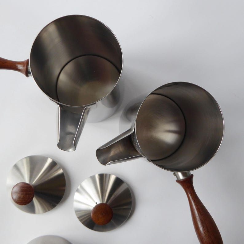 May include: Two stainless steel coffee pots with wooden handles and two matching lids. The pots have a spout and a rounded body.