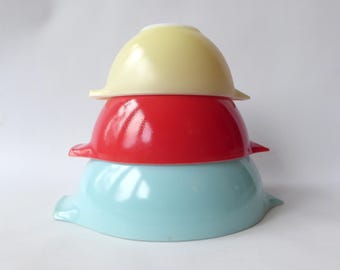 May include: Three vintage mixing bowls stacked. The top bowl is pale yellow, the middle bowl is red, and the bottom bowl is light blue. Each bowl has a flared rim. A retro kitchenware item.