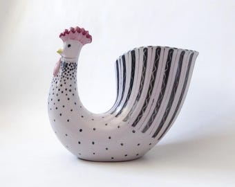 May include: A ceramic rooster-shaped vessel with a white base colour. The neck features black polka dots, a red comb, and a yellow beak. The body transitions into a wide, open tail section decorated with vertical black stripes and wavy lines.