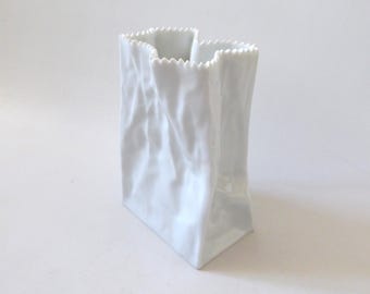 Könnte beinhalten: Eine weiße Keramikvase in Form einer zerknitterten Papiertüte. Das Objekt hat eine glänzende Oberfläche, einen rechteckigen Boden und einen gezackten oberen Rand, der die Optik einer aufgerissenen Papieröffnung mit tiefen Falten imitiert.