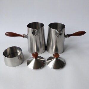 May include: Set of two stainless steel creamer pitchers with wooden handles and lids. The pitchers have a polished finish and are designed for serving cream or milk.
