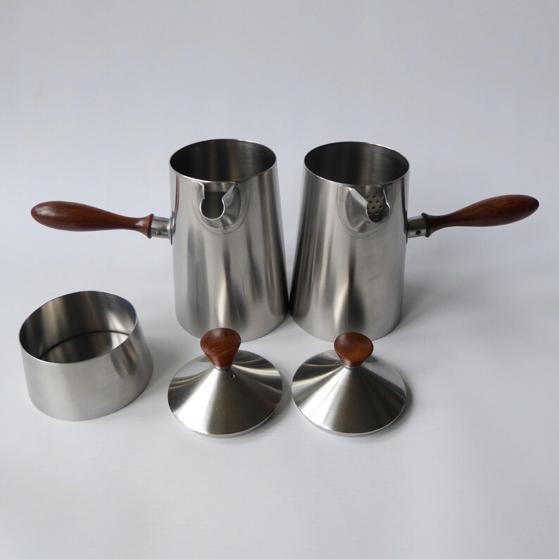 May include: Set of two stainless steel creamer pitchers with wooden handles and lids. The pitchers have a polished finish and are designed for serving cream or milk.