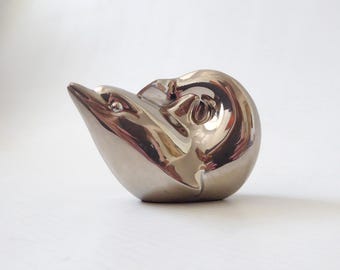 May include: A polished, metallic silver-toned sculpture of a dolphin in a curved, leaping posture. The surface is highly reflective, showing smooth, rounded contours and a streamlined form that captures the essence of the aquatic mammal.