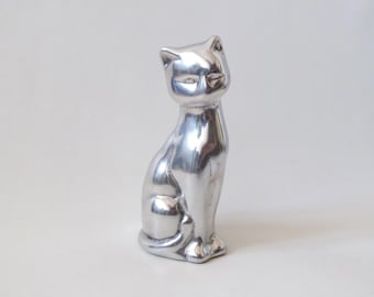 Könnte beinhalten: Eine polierte silberfarbene Metallfigur einer sitzenden Katze. Die Skulptur hat eine glatte, reflektierende Oberfläche mit stilisierten, vereinfachten Gesichtszügen, spitzen Ohren und einem Schwanz, der ordentlich um die Basis des Katzenkörpers gewickelt ist.