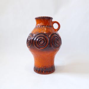 May include: A ceramic vase with a handle, in shades of orange and brown. The vase has a textured surface and is decorated with three dark brown spiral designs. The rim and base have a darker, textured finish.