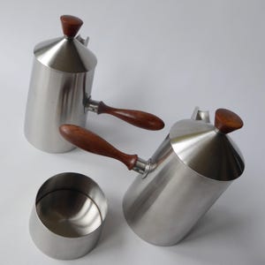 May include: Two silver pitchers with wooden handles and lids. The pitchers have a conical shape and are designed for pouring. A small silver bowl is also included.