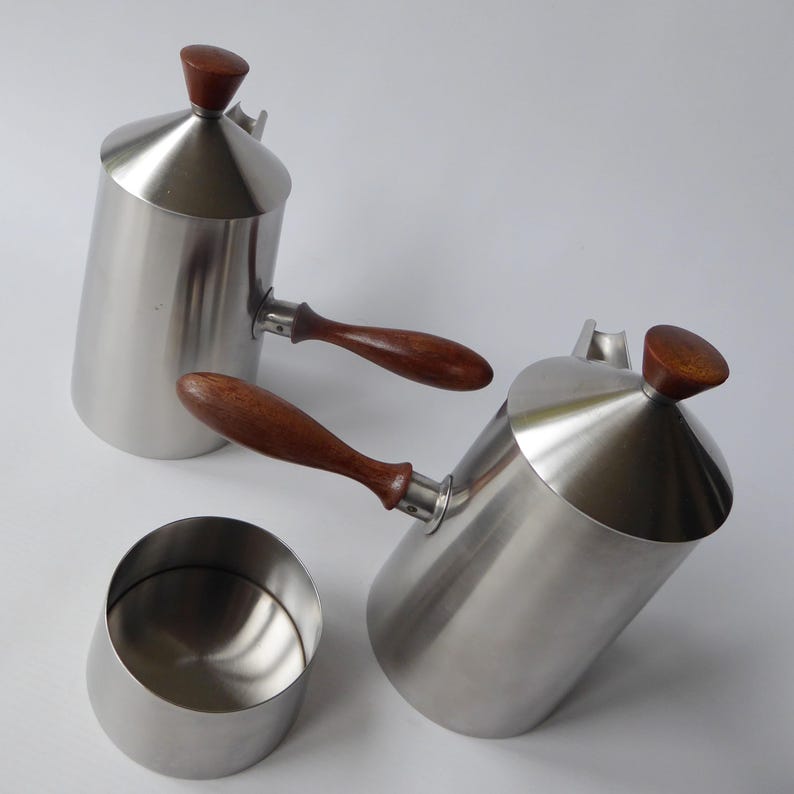 May include: Two silver pitchers with wooden handles and lids. The pitchers have a conical shape and are designed for pouring. A small silver bowl is also included.