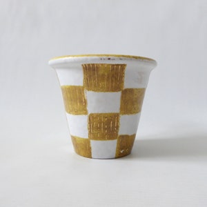 May include: A white ceramic planter with a checkered pattern in gold. The planter has a wide mouth and a narrow base.
