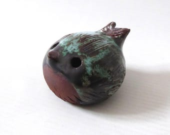 Könnte beinhalten: Eine runde Keramik-Vogelfigur mit einer dunkelbraunen und türkisfarbenen Glasur. Die Skulptur hat einen glatten, rotbraunen Brustbereich, einen kleinen spitzen Schnabel und zwei kreisförmige Augenöffnungen. Die Oberfläche weist texturierte Details auf.
